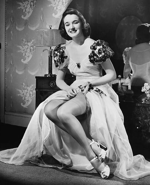 Woman in evening gown putting on stockings at vanity table (B&W), portrait