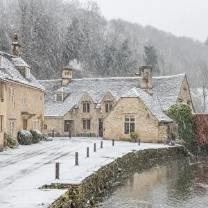 Snow covered houses by By Brook in Castle Combe with a dog enjoying a paddle, Wiltshire